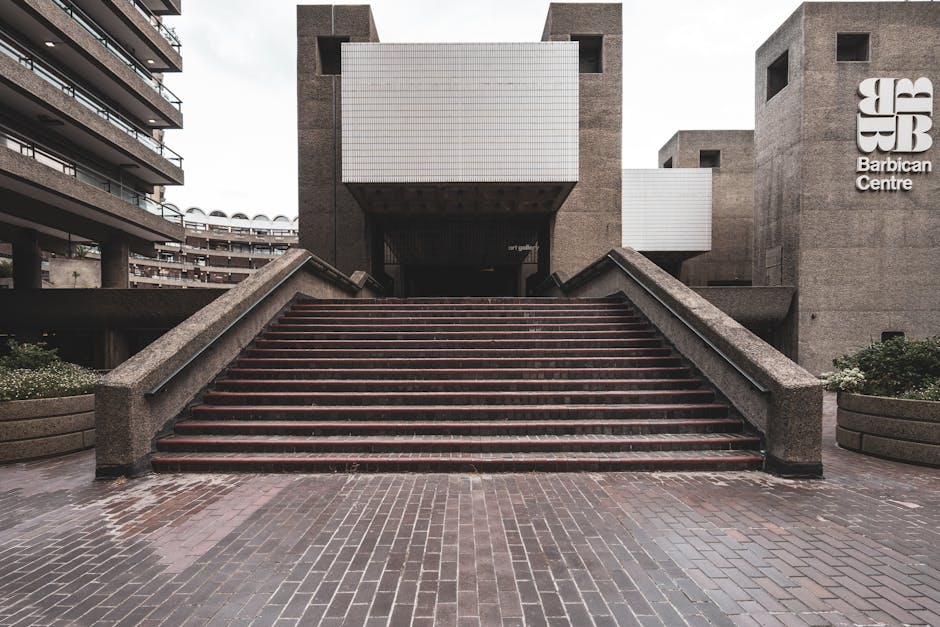 Interior view of a spacious, carpeted area inside the Barbican Centre showcasing a seating zone with chairs and tables, illuminated by warm lighting from wall-mounted fixtures. The carpet appears clea